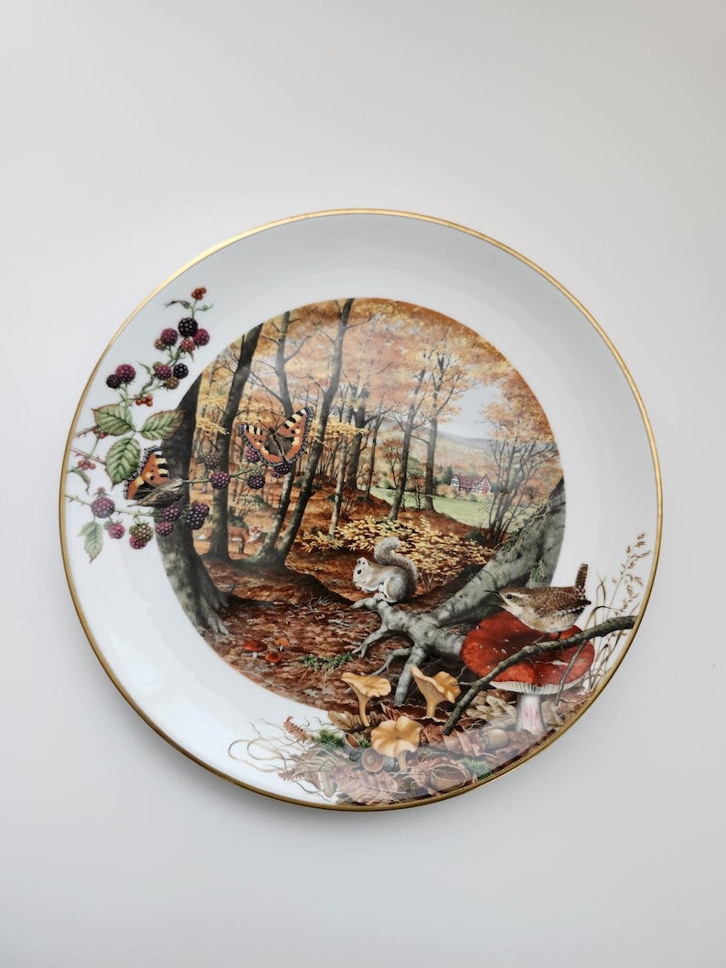 May include: A decorative white plate with a gold rim featuring a detailed forest scene. The scene includes a squirrel, mushrooms, a bird, and butterflies. The plate also has a branch with blackberries.