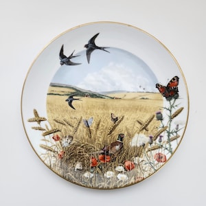 May include: A decorative plate with a gold rim featuring a pastoral scene. The design includes swallows in flight, butterflies, wheat, poppies, and a pheasant. The plate has a white background.