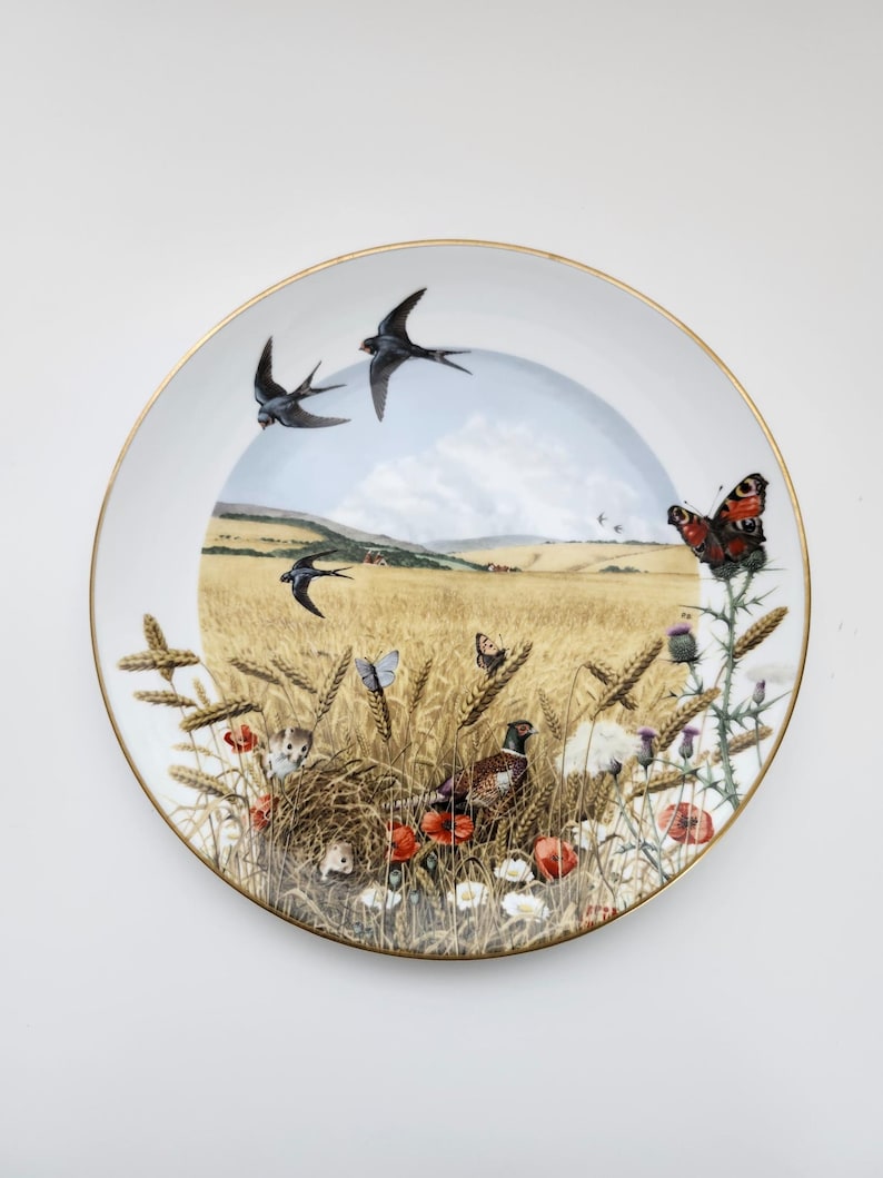 May include: A decorative plate with a gold rim featuring a pastoral scene. The design includes swallows in flight, butterflies, wheat, poppies, and a pheasant. The plate has a white background.
