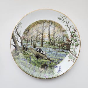May include: A white decorative plate with a gold rim featuring a detailed forest scene. The scene includes trees, bluebells, a badger, a hedgehog, and a bird. The plate's design is a nature-themed illustration.