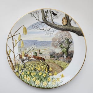 May include: A decorative ceramic plate with a gold rim. The plate features a detailed landscape scene with a church, a path, and yellow daffodils. Birds perch on a tree branch, and an owl is also visible.