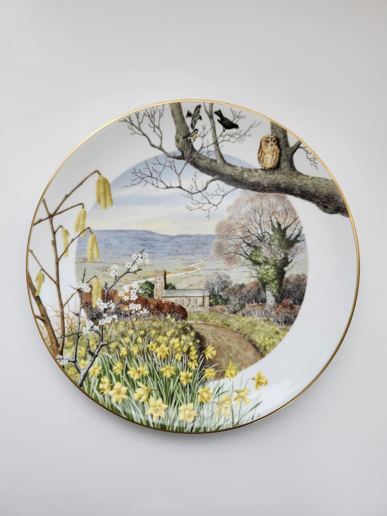 May include: A decorative ceramic plate with a gold rim. The plate features a detailed landscape scene with a church, a path, and yellow daffodils. Birds perch on a tree branch, and an owl is also visible.