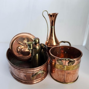 May include: A collection of copper and brass kitchenware including a round lidded container with an apple motif, a tall slender pitcher with a curved handle, a wide shallow basin with side handles, and a cylindrical bucket with brass accents.