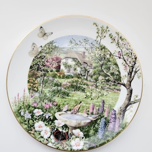May include: A decorative plate featuring a detailed garden scene with a cottage, flowers, and butterflies. The plate has a gold rim and a serene, colorful landscape design. Birds are perched on a birdbath. The plate is white.