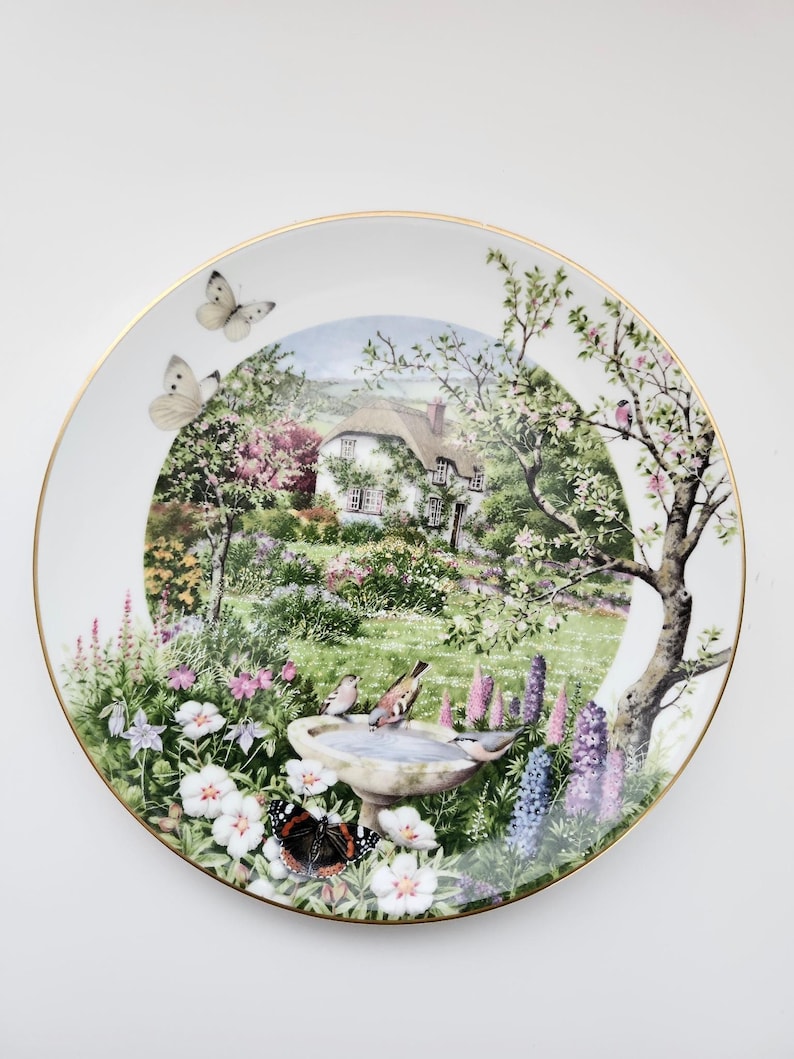 May include: A decorative plate featuring a detailed garden scene with a cottage, flowers, and butterflies. The plate has a gold rim and a serene, colorful landscape design. Birds are perched on a birdbath. The plate is white.