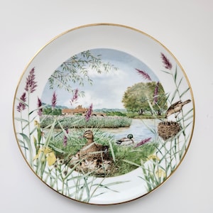 May include: A decorative porcelain plate with a gold rim. The plate features a detailed painting of a lakeside scene with ducks, birds, and reeds. The artwork includes a house in the background and a blue sky.