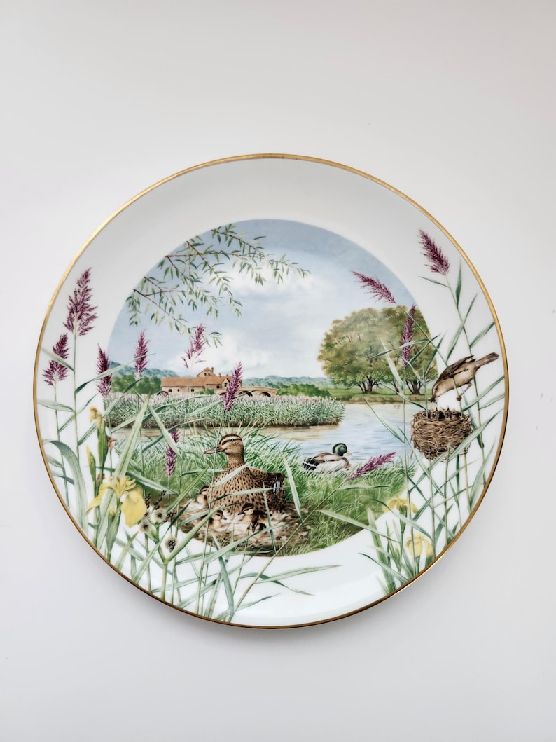 May include: A decorative porcelain plate with a gold rim. The plate features a detailed painting of a lakeside scene with ducks, birds, and reeds. The artwork includes a house in the background and a blue sky.