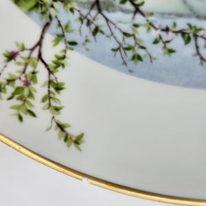 May include: White porcelain plate with a gold rim, featuring a painted scene of a tree branch with green leaves and pink blossoms. The background has a light blue sky and a glimpse of a building.
