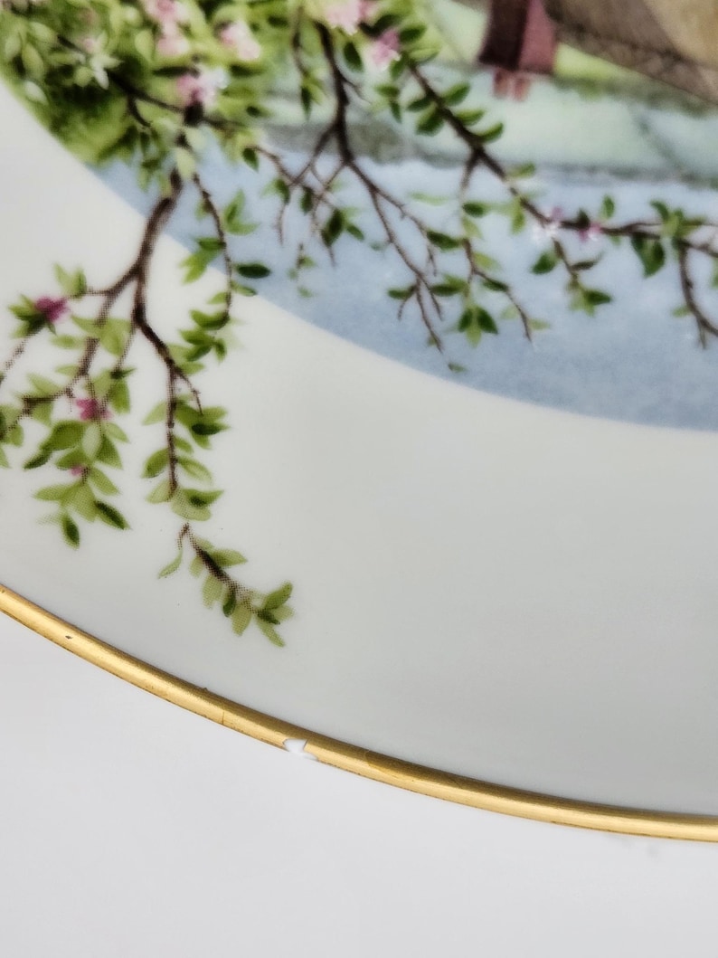 May include: White porcelain plate with a gold rim, featuring a painted scene of a tree branch with green leaves and pink blossoms. The background has a light blue sky and a glimpse of a building.
