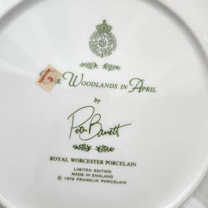May include: White porcelain plate with green text that reads "The Woodlands in April" by Peter Barrett. The plate is a limited edition from Royal Worcester Porcelain, made in England in 1979. A small sticker reads "Inspected 3E".