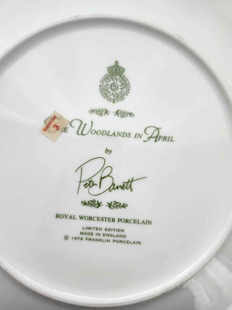 May include: White porcelain plate with green text that reads "The Woodlands in April" by Peter Barrett. The plate is a limited edition from Royal Worcester Porcelain, made in England in 1979. A small sticker reads "Inspected 3E".