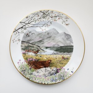 May include: A decorative plate with a gold rim features a scenic landscape with mountains, a lake, and wildlife. The plate showcases a detailed painting of deer, a bird, and wildflowers. The plate is white with a gold trim.