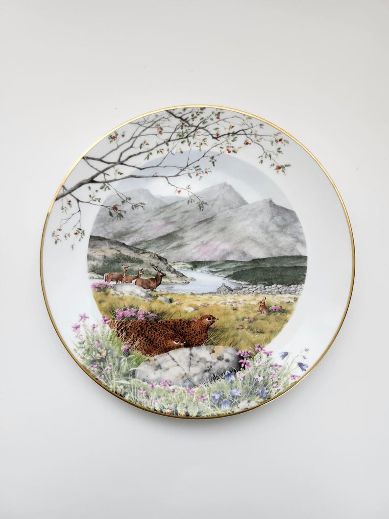 May include: A decorative plate with a gold rim features a scenic landscape with mountains, a lake, and wildlife. The plate showcases a detailed painting of deer, a bird, and wildflowers. The plate is white with a gold trim.