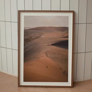 May include: An aerial view of a vast desert landscape with a single figure walking across a sand dune. The sand is a warm orange color and the sky is a pale blue. The image is framed in a wooden frame.
