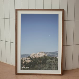 May include: A framed photograph of a cityscape with a hilltop view of a building complex against a blue sky with a small moon visible. The image is framed in a brown wood frame.