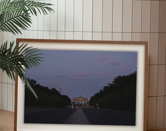 Puede incluir: Una fotografía horizontal enmarcada que muestra la Puerta de Brandeburgo en Berlín al atardecer. La imagen presenta el monumento iluminado al final de una carretera oscura bajo un cielo crepuscular púrpura y rosa, con la torre de televisión a lo lejos.