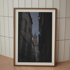 Könnte beinhalten: Eine enge, steinerne Gasse in einer europäischen Stadt mit einem hohen, steinernen Kirchturm am Ende. Die Gasse ist von alten, steinernen Gebäuden gesäumt.