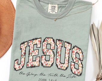 Peut inclure: T-shirt vert sauge avec le mot "JESUS" dans un motif floral. Le texte "the Way, the truth, the Life" et "JOHN 14:6" sont imprimés en dessous. Un collier doré avec un pendentif en forme d'étoile est sur le t-shirt.