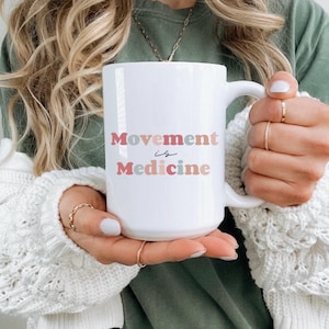 Puede incluir: Taza de cer&aacute;mica blanca con el texto "Movement is Medicine" en letras rosas, azules y melocot&oacute;n. La taza tiene un asa grande y la sostiene una persona que lleva anillos y un su&eacute;ter blanco.