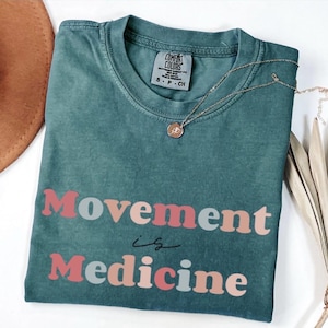 Puede incluir: Una camiseta Comfort Colors de color verde azulado con la frase "Movement is Medicine" en una fuente retro colorida. Un collar de oro con un colgante descansa sobre la camisa. La camisa est&aacute; doblada y se muestra sobre una superficie blanca.