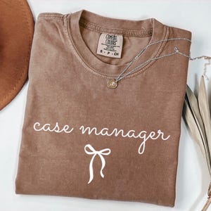 May include: A brown Comfort Colors t-shirt with the words "case manager" in white script and a bow graphic. A silver necklace with a pendant rests on the shirt. The shirt is folded.
