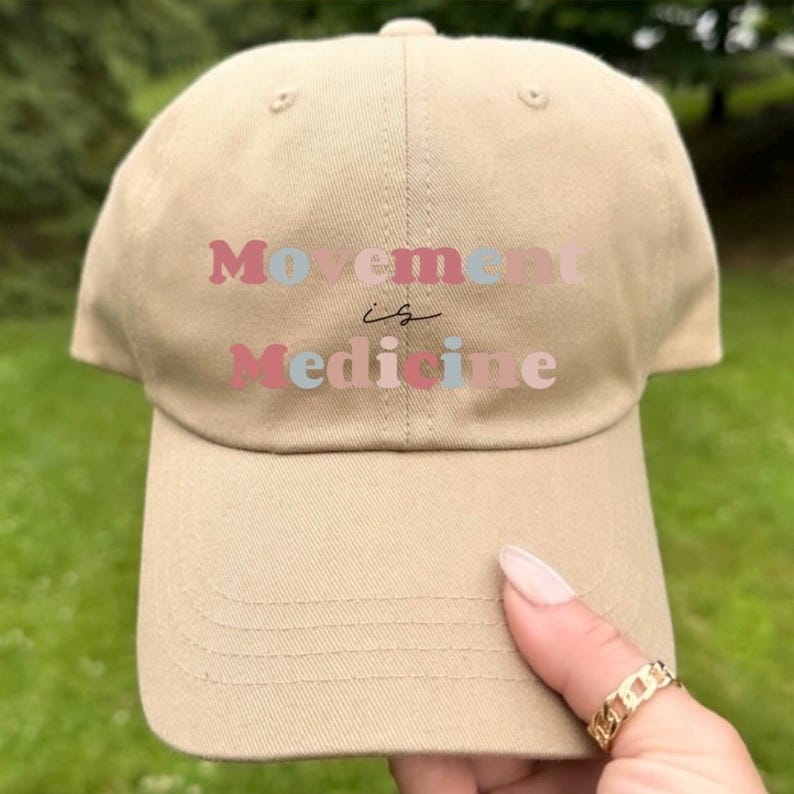 Puede incluir: Gorra de b&eacute;isbol beige con la frase "Movement is Medicine" en letras rosas, azules y rojas. La gorra est&aacute; hecha de tela suave y tiene una visera curva. Una persona sostiene la gorra.