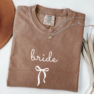 May include: A brown Comfort Colors t-shirt with the word "bride" in white script and a white bow graphic. A silver necklace with a small pendant is draped across the shirt. The shirt is folded and ready to wear.