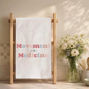 Puede incluir: Toalla blanca de tejido de gofre con el texto "Movement is Medicine" en letras rosas y azules. La toalla se muestra en un toallero de madera clara. Un jarr&oacute;n de flores blancas est&aacute; cerca.
