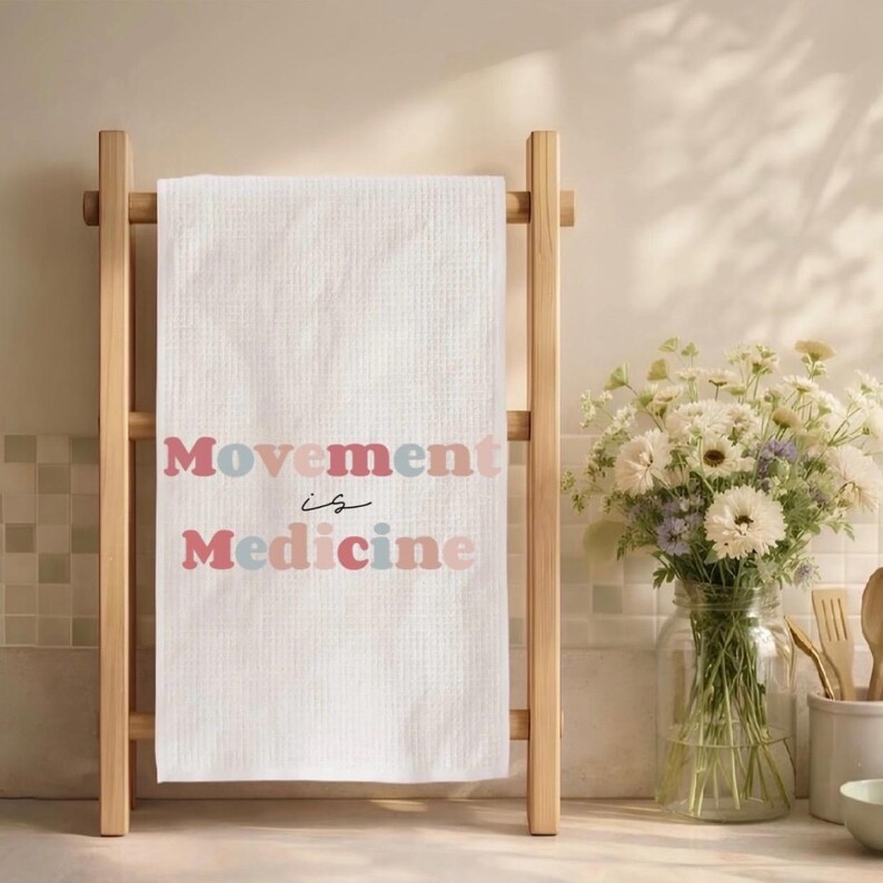 Puede incluir: Toalla blanca de tejido de gofre con el texto "Movement is Medicine" en letras rosas y azules. La toalla se muestra en un toallero de madera clara. Un jarr&oacute;n de flores blancas est&aacute; cerca.