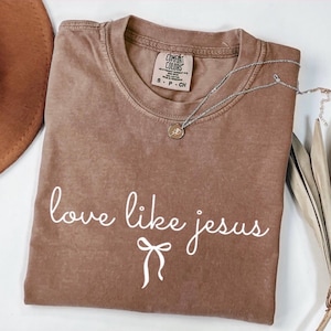 May include: A brown Comfort Colors t-shirt with the phrase "love like jesus" in white script. A silver necklace with a small pendant rests on the shirt. A brown hat is partially visible in the upper left corner.