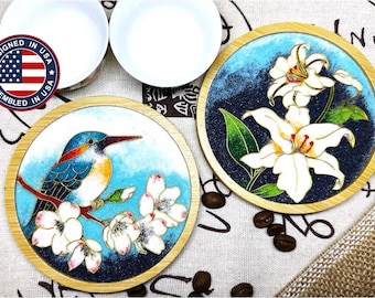 Può includere: Due sottobicchieri rotondi in legno con disegni dipinti. Un sottobicchiere presenta un uccello blu appollaiato su un ramo con fiori bianchi. L'altro sottobicchiere presenta un giglio bianco con foglie verdi su uno sfondo blu.