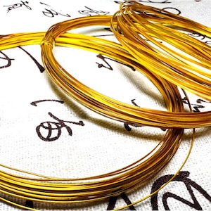 May include: Several coils of shiny, golden-colored wire are arranged on a white fabric with black calligraphy. The wire is tightly wound into circular shapes, suggesting its use in jewelry making or crafts. The lighting highlights the metallic sheen.