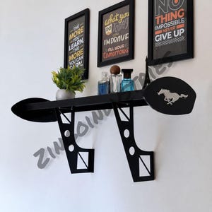 May include: A black wall-mounted shelf shaped like a car spoiler with two support brackets. The right side features a silver horse emblem. On the shelf are a small potted green plant, three glass perfume bottles, and a miniature Eiffel Tower model.