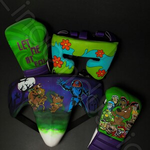 Könnte beinhalten: Ein Boxausrüstung-Set mit Cartoon-Charakter-Designs. Das Set enthält grüne und lila Boxhandschuhe mit dem Text "LET ME AT EM" und Charakterillustrationen. Ein passender Kopfschutz und Tiefschutz sind ebenfalls enthalten.