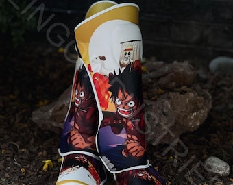 Könnte beinhalten: Ein Paar weiße und goldene Schienbeinschoner mit einem farbenfrohen Anime-Charakterdesign. Der Charakter trägt ein rotes Shirt und eine schwarze Hose. Die Schienbeinschoner haben einen weißen und goldenen Besatz.
