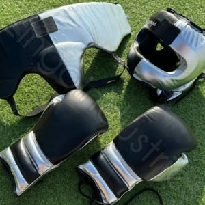 Op de afbeelding: Een set boksapparatuur, inclusief zwarte en zilveren bokshandschoenen, een hoofdbeschermer en een borstbeschermer. De handschoenen hebben zilveren accenten en zijn van leer. De hoofdbescherming en borstbescherming zijn zwart en zilver.