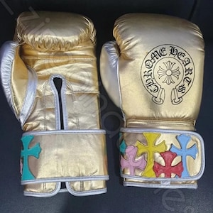 Könnte beinhalten: Goldene Boxhandschuhe mit silbernen Akzenten. Auf einem Handschuh steht "CHROME HEARTS" mit einem Kreuz-Design. Die Handgelenksriemen zeigen bunte Kreuzapplikationen. Boxhandschuhe.