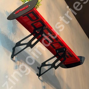 Könnte beinhalten: Ein roter Heckspoiler für Autos, der an einer Wand montiert ist, mit schwarzen Halterungen und Endplatten. Die Oberseite zeigt den roten Schriftzug GT-RS, während der rote Hauptteil große schwarze Buchstaben aufweist. Eine kleine Pflanze steht darüber.