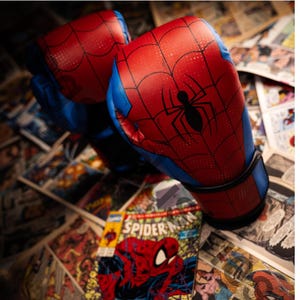 Pode incluir: Par de luvas de boxe vermelhas e azuis com design de teia de aranha e um emblema de aranha preta. As luvas estão sobre um fundo de páginas de banda desenhada, com uma banda desenhada do Homem-Aranha visível.
