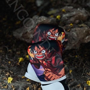 Puede incluir: Un par de guantes de boxeo blancos con correas de muñeca negras y un diseño de personaje de anime colorido que presenta un personaje sonriente con cabello rojo y una camisa roja. El personaje lleva un sombrero de paja y tiene un parche en el ojo negro.