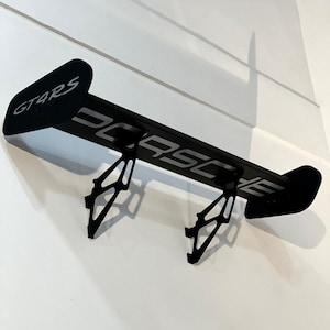 May include: Black metal car spoiler with the text "GT4RS" and "PORSCHE" in white. The spoiler is mounted on a white wall. The spoiler has two support brackets.