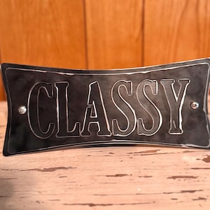 Może przedstawiać: Ciemnoszary metalowy znak z wyciętym słowem "CLASSY" w pogrubionej czcionce szeryfowej. Znak ma zakrzywiony kształt i jest zamontowany za pomocą dwóch małych srebrnych nitów. Tło to jasna drewniana powierzchnia.