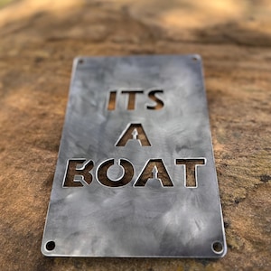 May include: A rectangular metal sign with the words "ITS A BOAT" cut out. The sign has a brushed metal finish and four holes in the corners. The sign is resting on a brown, textured surface.