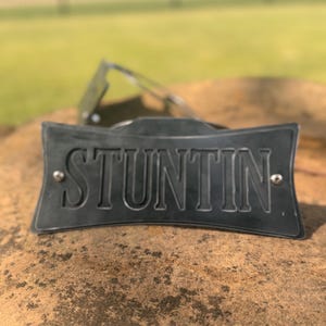 May include: A dark gray metal plate with the word "STUNTIN" embossed in capital letters. The plate is rectangular with a curved top and bottom. Two silver rivets are visible. Sunglasses are in the background.