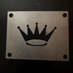 May include: A silver metal square with four holes around the edges and a black crown cut out in the center.