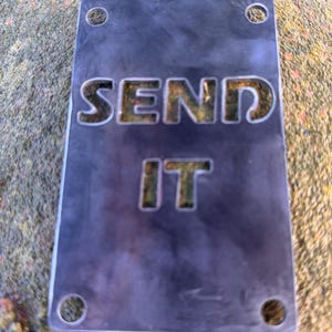 May include: A rectangular, dark gray metal sign with the words "SEND IT" cut out. The sign has four holes, one in each corner. The sign is sitting on a textured, light brown surface.