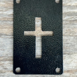 May include: A black metal plaque with a textured surface and a cut-out cross design. The rectangular plaque has rounded corners and four holes, one in each corner. The cross is centered on the plaque.