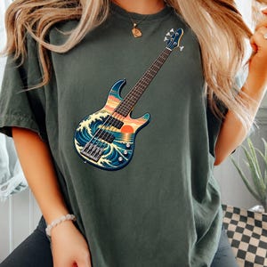 May include: A dark olive green t-shirt featuring a graphic print of an electric bass guitar. The guitar body displays a stylized illustration of a large ocean wave, a sunset, and a calm sea. The design includes a fretboard, strings, and knobs.