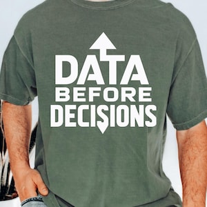 May include: A sage green short-sleeved t-shirt featuring the white text "DATA BEFORE DECISIONS" printed on the chest. The word "DATA" is positioned above the word "BEFORE" and "DECISIONS," with a white upward arrow above the word "DATA."