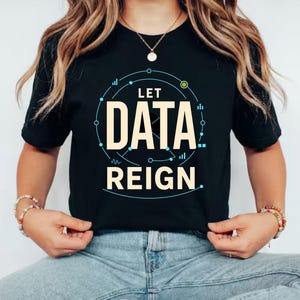 May include: A black short-sleeved t-shirt featuring the text LET DATA REIGN in large beige capital letters. The design includes a thin blue circular graphic with small geometric lines and dots surrounding the text, centred on the chest.